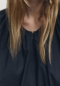 Svart blus med rund halsringning och kort dragkedja. Tyget verkar lätt och har en slät yta med en lätt glans.