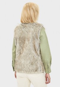 Veste en fausse fourrure beige et gris clair, avec un fini texturé et duveteux. Portée par-dessus une chemise vert clair, à manches longues et avec des poignets boutonnés.