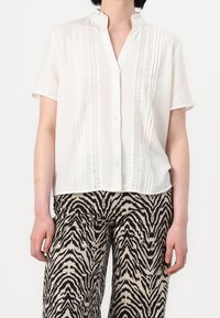 Žena v bílém krátkém rukávu s knoflíky, který má krajkové detaily, a v kalhotách se zebřím vzorem v černé a béžové barvě.