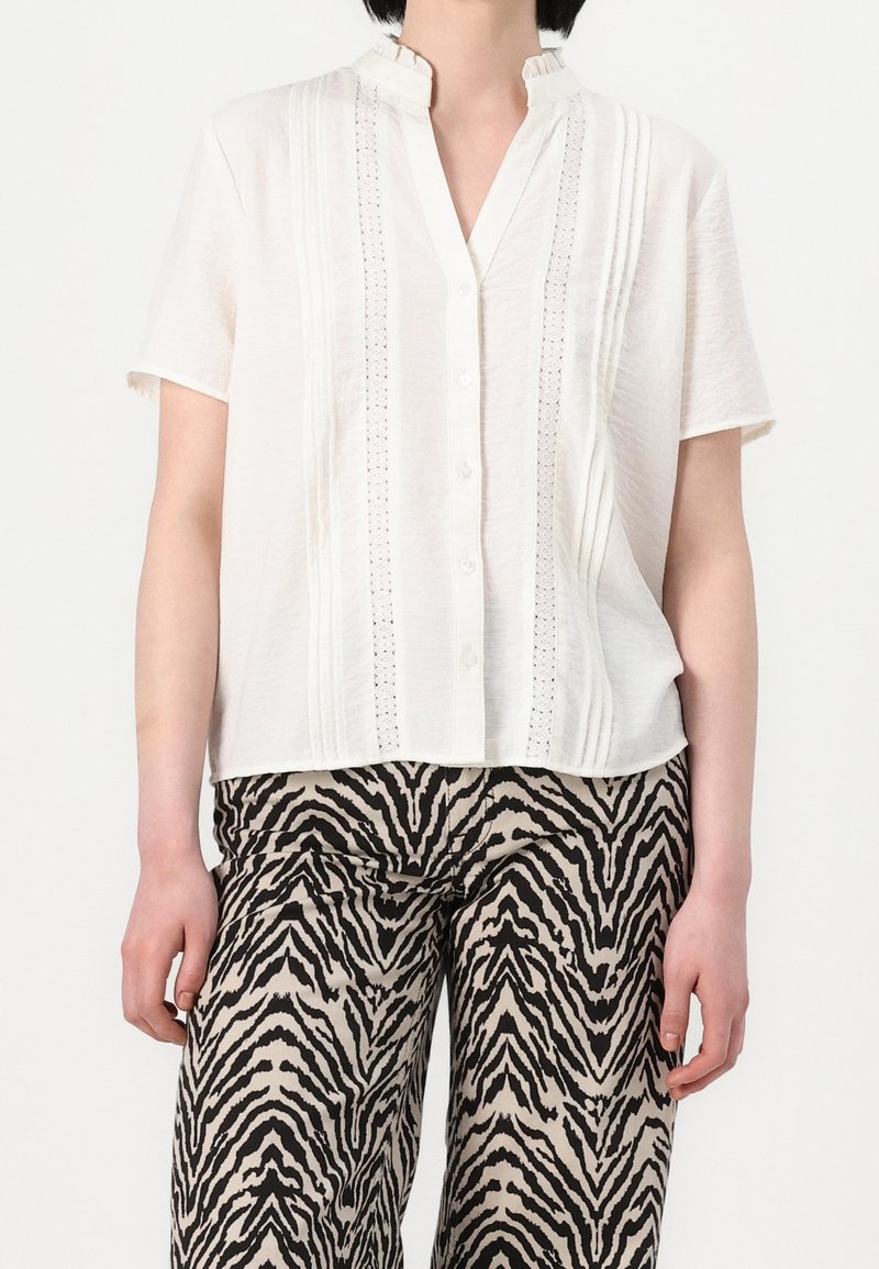 Žena v bílém krátkém rukávu s knoflíky, který má krajkové detaily, a v kalhotách se zebřím vzorem v černé a béžové barvě.