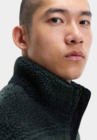 Fleece'owy kurtka w ciemnej zieleni, z wysokim kołnierzem, teksturowaną powierzchnią i kontrastującym czarnym suwakiem. Gładkie krawędzie, miękkie w dotyku.