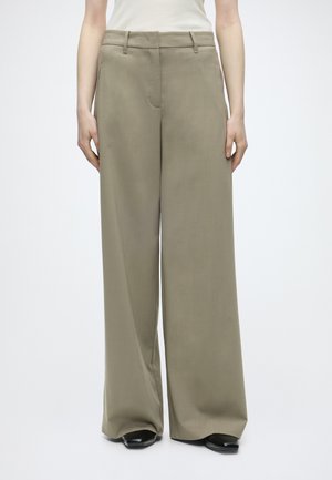 Γυναίκα που φοράει μπεζ παντελόνες με ψηλή μέση και φαρδιά μπατζάκια με θηλιές για ζώνη και μαύρα μυτερά παπούτσια, στέκεται μπροστά από ένα απλό φόντο.