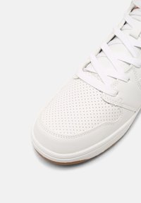 Bílé kožené tenisky s perforovanou špičkou, bílými tkaničkami a gumovou podrážkou na jemně šedém pozadí.