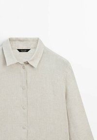 Chemise en lin beige clair à manches longues avec col et boutons sur le devant, montrée de la poitrine vers le haut sur un fond blanc.