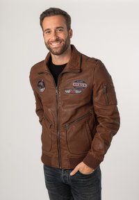 Braune Lederbomberjacke mit bestickten Patches, Reißverschlusstaschen und gerippten Bündchen. Sie verfügt über einen Frontreißverschluss und kontrastierende Texturen.