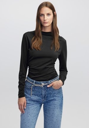 Schwarzes Langarmoberteil mit hohem Kragen, kombiniert mit hellblauen Jeans mit Nietenverzierungen und einem dekorativen Gürtel.