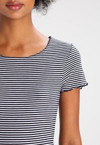 Czarny i biały t-shirt w paski z okrągłym dekoltem i krótkimi rękawami. Rękawy mają falbaniaste wykończenie, a materiał wydaje się prążkowany.