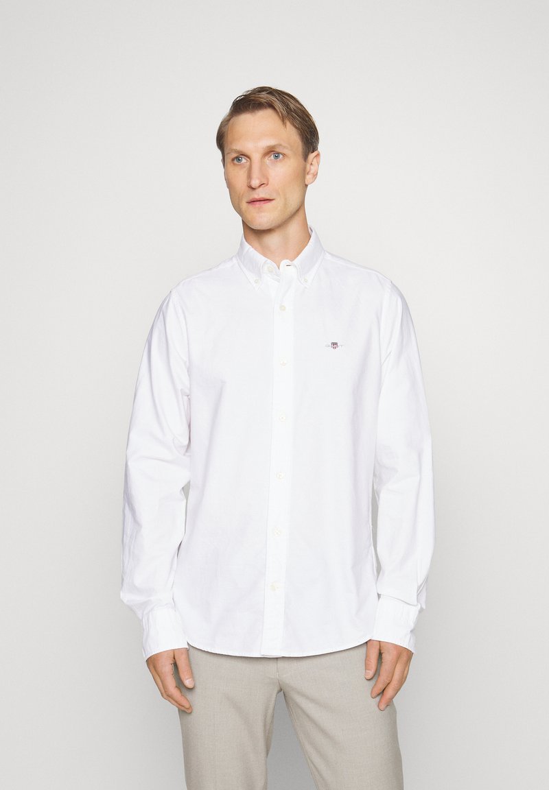 Homme portant une chemise blanche boutonnée et un pantalon beige, debout devant un fond clair uni, regardant légèrement sur le côté.