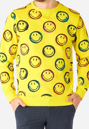 Mand iført en knaldgul sweatshirt dekoreret med flerfarvede smiley-ansigter, hænderne hviler i lommerne på mørke teksturerede bukser.