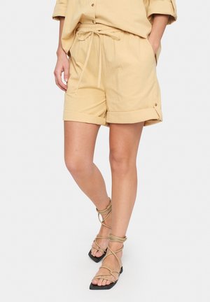 Beige linen shorts with an elastic waistband and drawstring, featuring rolled cuffs and side pockets, paired with strappy black sandals.