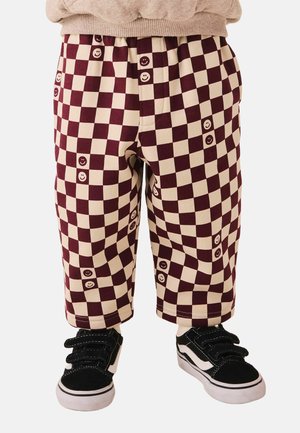 Child wearing wide-legged, beige and maroon checkered pants with smiley faces, black and white sneakers, and a beige sweater.