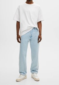 Camiseta de algodón blanca de manga corta, combinada con jeans de mezclilla azul claro y zapatillas blancas con suelas marrones. Ajuste casual y líneas limpias.