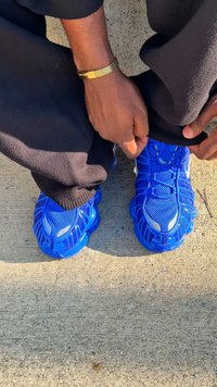 Ténis desportivos azuis com parte superior em malha texturizada, solas curvas e marcações notáveis. Pessoa ajusta calças pretas acima dos sapatos.