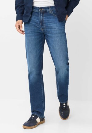 Uomo che indossa jeans blu medi a gamba dritta, sneakers blu navy con strisce bianche, camicia bianca e giacca blu navy, camminando con una mano in tasca.