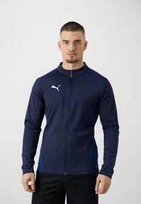Marine atletisk jakke med lynlås foran, ståkrave og hvidt logo. Har en tætsiddende design og kontrasterende blå sidepaneler.