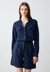 Camicia button-up in lino blu navy con due tasche sul petto e pantaloni corti coordinati, caratterizzati da una vestibilità rilassata e un design minimalista.