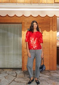 Haut en sequins rouges à manches courtes, associé à un jean gris déchiré. Chaussures noires slip-on et sac à main noir à imprimé léopard. Fond en panneaux de bois.