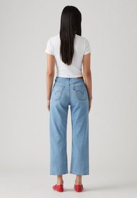 Femme aux longs cheveux foncés, debout de dos, portant un t-shirt court blanc, un jean large bleu clair et des chaussures plates rouges.