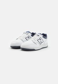 Hvide lædersneakers med marineblå detaljer, perforeret tå, flade snørebånd og gummisål. Har et fremtrædende "N" logo på siden.