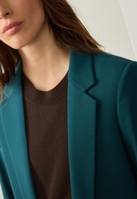 Teal blazer with structured lapels paired with a brown sweater. Blazer features a smooth texture; sweater has a ribbed crew neckline.