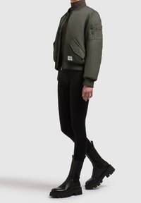 Personne portant une veste bomber vert olive, des leggings noirs et des bottes en cuir noires épaisses, debout sur un fond uni gris clair.