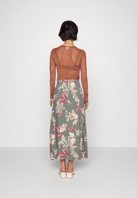 Haut en chiffon marron à manches longues associé à une jupe maxi florale verte ornée de fleurs roses et de papillons ; le mannequin porte des chaussures à talons carrés blanches.