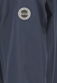Pantalons outdoor navy en tissu léger, dotés d'un patch logo rond gris "ZIGZAG" avec texte, affichant une texture lisse et mate.