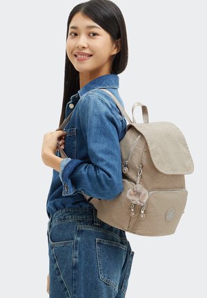 Ung kvinde i denimoutfit, der bærer en beige rygsæk med en abe nøglekæde, smilende og kigger over sin skulder.