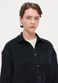 Svart cordfløyelskjorte med kragehals, med frontknappestenging og en myk, strukturert overflate.