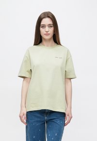 Hellgrünes Baumwoll-T-Shirt mit kurzen Ärmeln, rundem Ausschnitt und dezentem Markenschriftzug in dunkler Farbe auf der Brust. Kombiniert mit blauen, leicht zerrissenen Jeans.