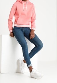 Sudadera rosa claro con bolsillo tipo canguro y cordones blancos, combinada con jeans skinny azul oscuro y zapatillas deportivas blancas.