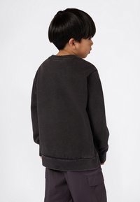 Ragazzo giovane con capelli corti e scuri, che indossa una felpa nera e pantaloni cargo scuri, girato di spalle su uno sfondo bianco uniforme.