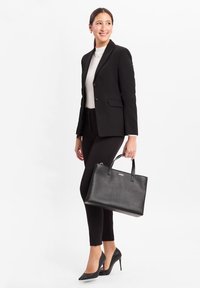 Borsa a tracolla in pelle nera con manici corti, superficie testurizzata e logo impresso. Il modello indossa un completo nero aderente e tacchi, in posa di profilo.
