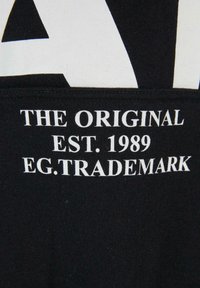 Texte blanc sur tissu noir indiquant "THE ORIGINAL EST. 1989 EG.TRADEMARK" en police serif en gras, centré sous un grand graphisme blanc.