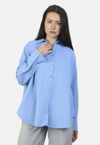 Camisa de manga larga azul claro con un diseño de botones, cuello y un leve brillo. Presenta un corte relajado y tiene un dobladillo curvado.