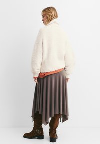 Plyšová, krémově zbarvená bunda s vysokým límcem, vrstvená přes oranžový top a splývavou taupe sukni. Hnědé semišové kozačky po kolena s sponami.