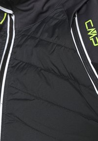 Veste isolée noire avec des sections matelassées, des accents blancs et un logo vert. Des poches zippées sont visibles sur les côtés.