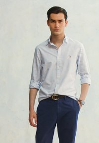 Camisa de botão riscada em azul claro e branco, combinada com calças azul-marinho. Apresenta mangas enroladas e um logótipo subtil no peito.