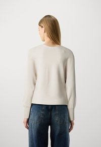 Light beige knitted sweater with ribbed texture; features long sleeves and a round neckline, paired with high-waisted blue denim jeans.