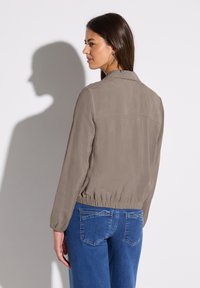 Veste zippée marron clair avec taille élastique et manches longues, associée à un jean en denim bleu. Texture lisse, design minimal.