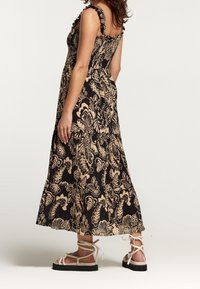 Black floral maxi dress with beige prints, smocked bodice, ruffled straps, and paired with light-colored strappy sandals on a flat sole.