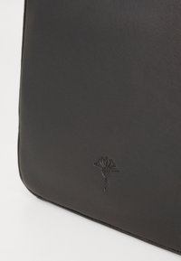 Glatt svart skinnveske hjørne med en subtil preget logo av en stilisert blomst eller palmeblad. Minimalistisk design og teksturer er synlige.