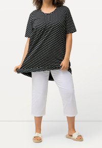 Schwarze Kurzarm-Tunika mit weißem geometrischen Muster, übergroßer Passform und abgerundetem Saum, kombiniert mit weißen Cropped-Hosen und leichten Sandalen.