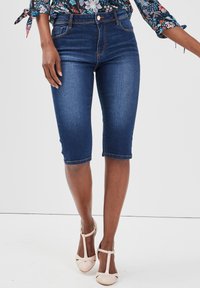 Shorts en denim bleu foncé avec un léger délavage, cinq poches et une coupe ajustée, associés à des chaussures à talons hauts rose clair.