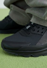 Chaussure de sport noire avec surface texturée, dotée d'une semelle épaisse et d'accents à motifs multiples, associée à un pantalon en velours côtelé de couleur claire.
