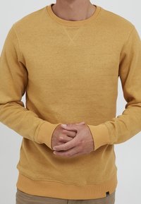 Żółta bluza musztardowa z okrągłym dekoltem, ściągaczami na rękawach i dole, miękką fakturą oraz małym logo w pobliżu klatki piersiowej.