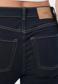 Un par de jeans de mezclilla azul oscuro con un diseño ajustado, que presentan costuras amarillas, dos bolsillos traseros y una etiqueta de cuero beige en la cintura.