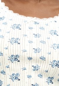 Κοντινή λήψη ενός λευκού πουκαμίσου με μπλε λουλουδάτα σχέδια, κάθετες διακεκομμένες γραμμές και λαιμόκοψη με δαντέλα και μικρό μπροστινό φιόγκο.