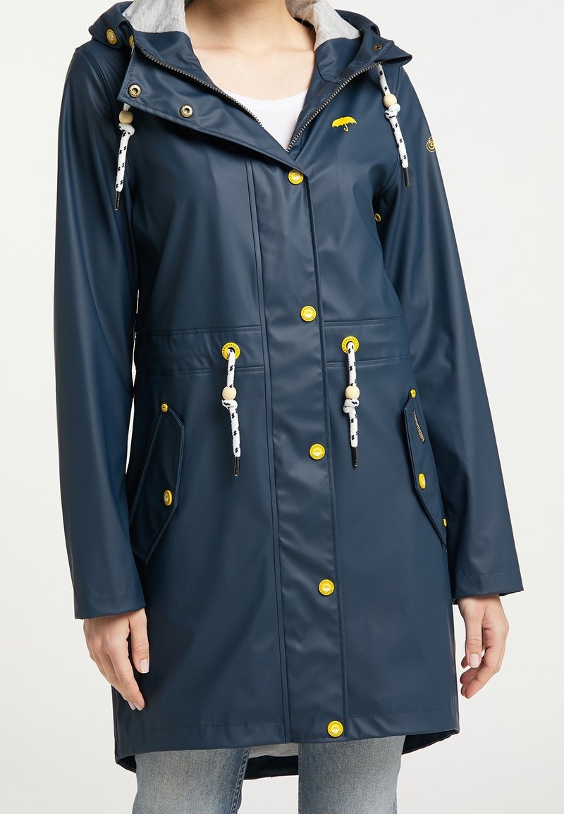 Femme portant un imperméable bleu marine avec des boutons jaunes, des cordons blancs avec des perles en bois, et des poches avant, sur un jean clair.