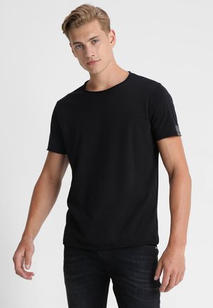 Camiseta negra de manga corta con cuello redondo, hecha de una tela suave, que presenta un sutil detalle de parche en la manga izquierda.